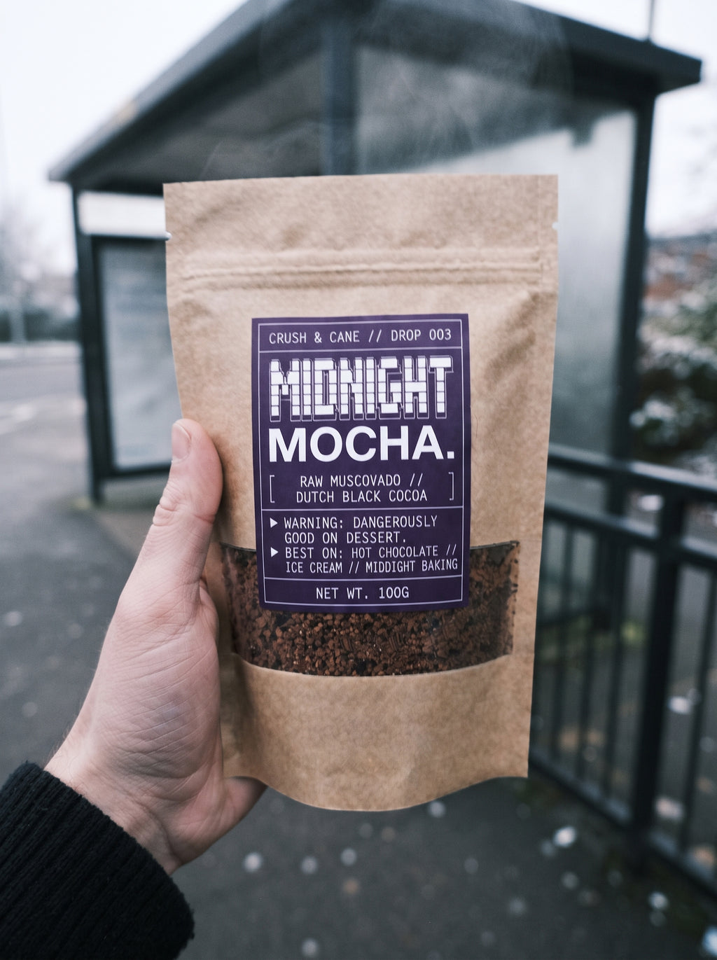 A hand holding a 100g kraft paper pouch of Midnight Mocha coffee. The pouch has a dark purple label with white text and a clear window showing the dark coffee granules inside.