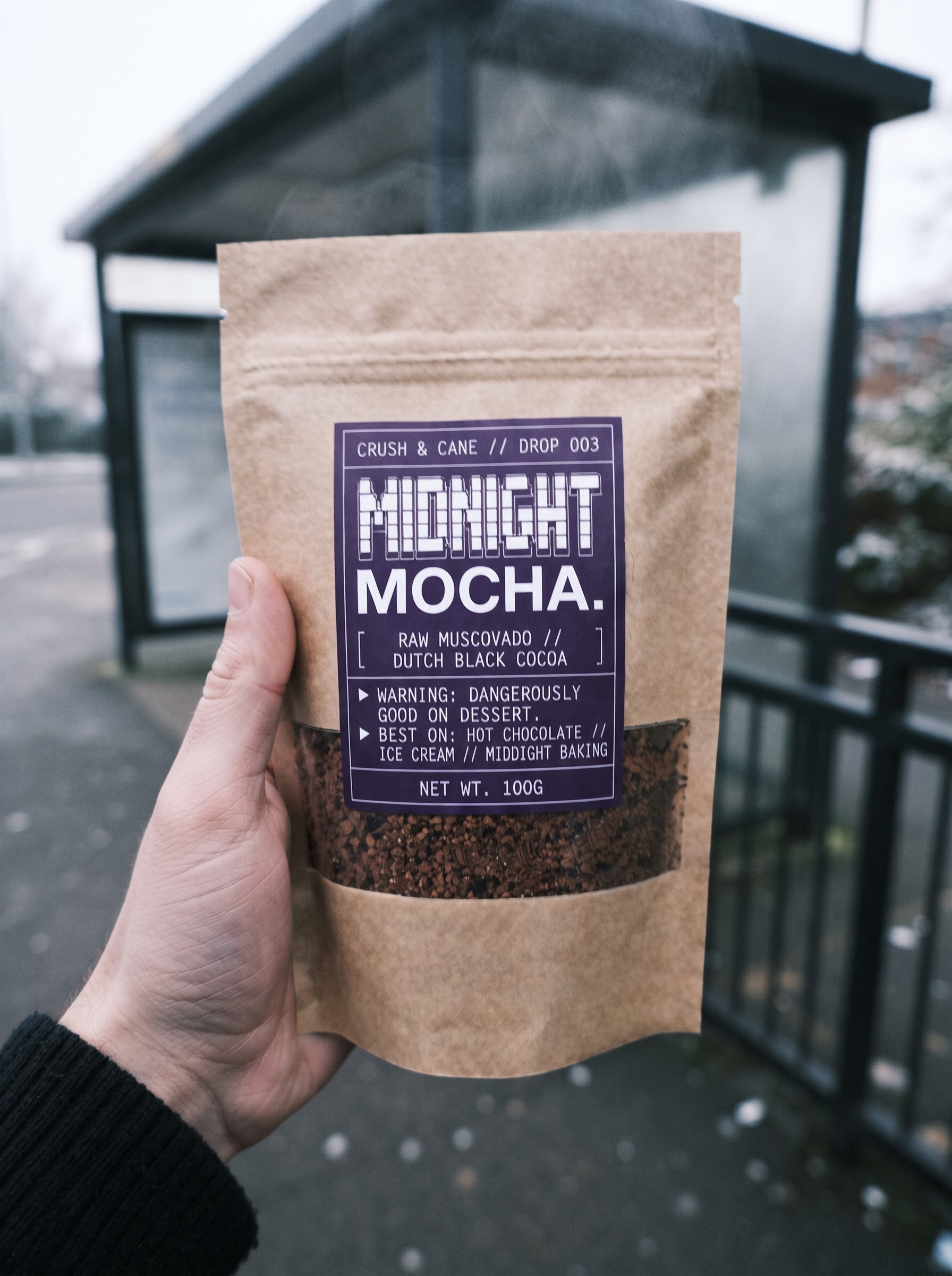 A hand holding a 100g kraft paper pouch of Midnight Mocha coffee. The pouch has a dark purple label with white text and a clear window showing the dark coffee granules inside.