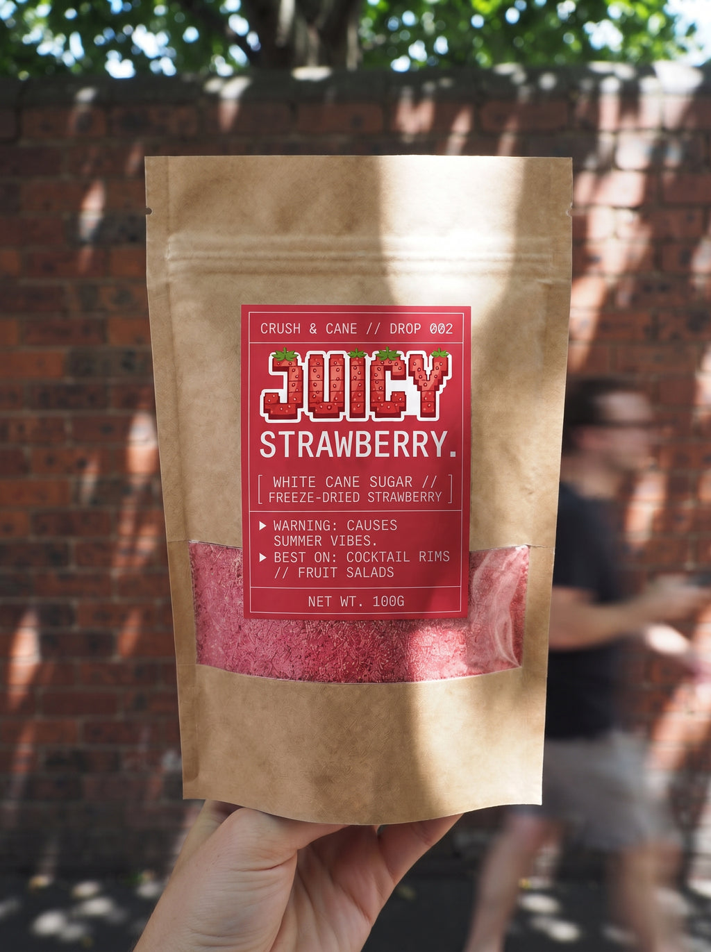 A hand holds a brown kraft stand-up pouch of Juicy Strawberry cane sugar in front of a blurred brick wall and outdoor background. The pouch has a red label with white text and a clear window showing pink-red sugar inside.