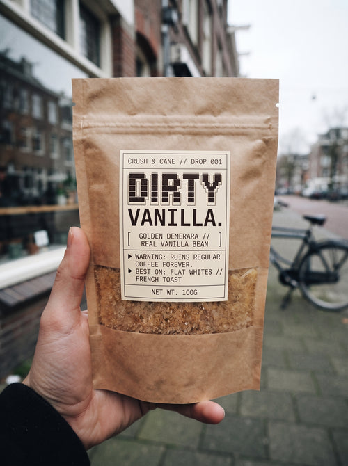 A hand holding a 100g kraft paper pouch of Dirty Vanilla coffee beans, with a blurred city street background.